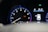 A car dashboard with a tachometer displaying RPM levels and warning lights illuminated, including oil and engine warnings. A digital screen shows the car in a parking mode with an open door and details like fuel range and comfort mode.
