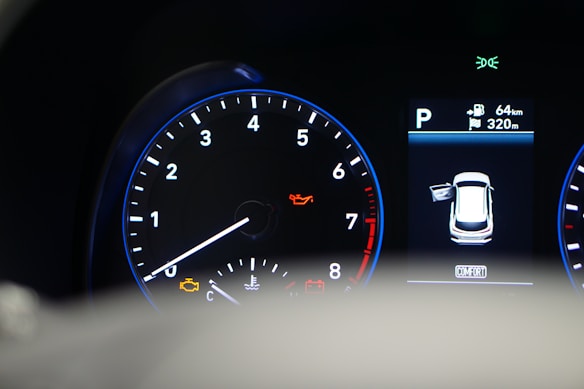 A car dashboard with a tachometer displaying RPM levels and warning lights illuminated, including oil and engine warnings. A digital screen shows the car in a parking mode with an open door and details like fuel range and comfort mode.