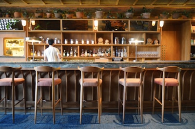 A cozy bar interior with wooden decor features a row of five high-backed chairs with cushioned seats along a marble countertop. The back wall is lined with an array of liquor bottles and glassware on wooden shelves. A person is standing behind the bar, facing away, serving or preparing drinks. Potted plants decorate the top shelves, adding a touch of greenery.