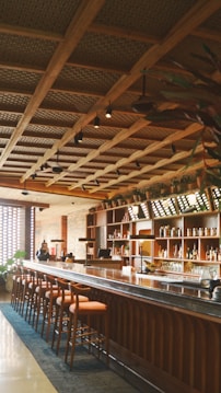 A stylish bar interior featuring a long wooden counter and several high chairs. The ceiling has a geometric grid design with hanging lights. Shelves behind the bar are filled with various bottles. There are potted plants adding a natural touch to the space.