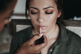 A person is having makeup applied to their lips, with closed eyes and a calm expression. The focus is on the lips, with a hand applying a light-colored lipstick or gloss.