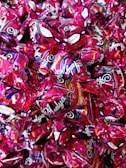 A collection of wrapped candies with shiny, metallic pink wrappers featuring decorative patterns and branding text. The candies are tightly packed together, creating a colorful and glossy surface.
