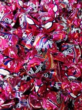 A collection of wrapped candies with shiny, metallic pink wrappers featuring decorative patterns and branding text. The candies are tightly packed together, creating a colorful and glossy surface.