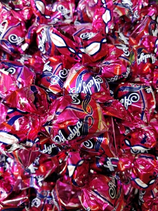 A collection of wrapped candies with shiny, metallic pink wrappers featuring decorative patterns and branding text. The candies are tightly packed together, creating a colorful and glossy surface.