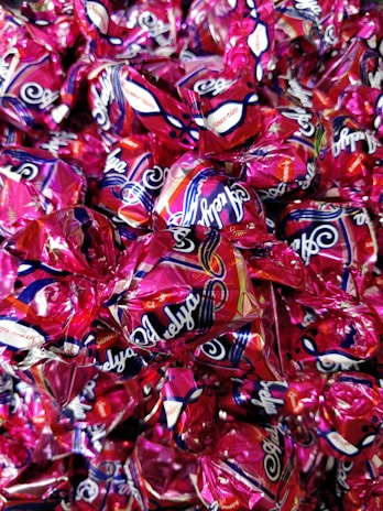 A collection of wrapped candies with shiny, metallic pink wrappers featuring decorative patterns and branding text. The candies are tightly packed together, creating a colorful and glossy surface.