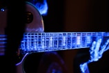 A close-up view of an electric guitar being played, with a focus on the fretboard and fingers. The image is illuminated with a blue and purple light, creating a luminous, almost ethereal effect on the guitar and the player's hands.