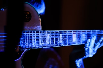 A close-up view of an electric guitar being played, with a focus on the fretboard and fingers. The image is illuminated with a blue and purple light, creating a luminous, almost ethereal effect on the guitar and the player's hands.