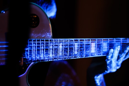 A close-up view of an electric guitar being played, with a focus on the fretboard and fingers. The image is illuminated with a blue and purple light, creating a luminous, almost ethereal effect on the guitar and the player's hands.