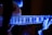 A close-up view of an electric guitar being played, with a focus on the fretboard and fingers. The image is illuminated with a blue and purple light, creating a luminous, almost ethereal effect on the guitar and the player's hands.