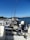 A row of fishing boats equipped with multiple fishing rods and tackle are docked by the water. The scene is set against a backdrop of clear blue skies and distant mountains. The water appears calm, reflecting the serene weather conditions.