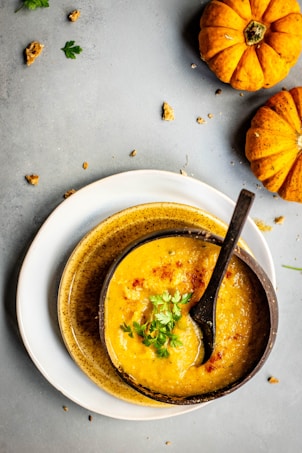 A bowl of creamy soup garnished with herbs is placed on a stack of ceramic plates. Whole small pumpkins and bread crumbs are scattered artistically on a gray surface, adding an autumnal touch.