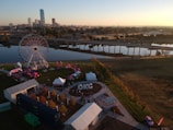 An aerial view of a park area with a large Ferris wheel near a body of water. The landscape includes several tents, likely indicating a festival or event, and the word 'OKC' is prominently displayed on the ground. Tall city buildings stand in the background, and it's nearing sunset.