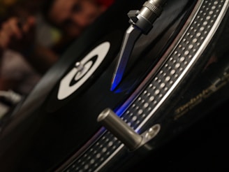 A close-up view of a vinyl record player, focusing on the needle and turntable. The record is spinning under the stylus, which is delicately placed on its grooves, emitting a blue light. The image depicts a classic DJ setup with a sense of motion.