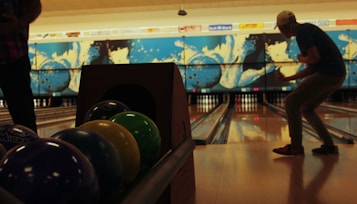 A bowling alley with colorful bowling balls lined up on a rack. The background features vibrant murals of bowling balls and pins. A person in a cap is captured in a dynamic pose as they bowl, focused on the lane.