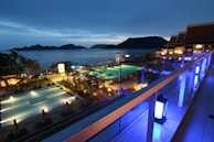 A nighttime scene overlooking a luxurious resort with multiple illuminated swimming pools. The view extends over a body of water with silhouetted islands in the distance. The area is adorned with palm trees and decorative lighting, creating a serene and inviting atmosphere.