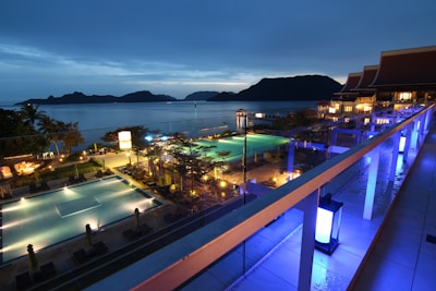 A nighttime scene overlooking a luxurious resort with multiple illuminated swimming pools. The view extends over a body of water with silhouetted islands in the distance. The area is adorned with palm trees and decorative lighting, creating a serene and inviting atmosphere.