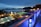A nighttime scene overlooking a luxurious resort with multiple illuminated swimming pools. The view extends over a body of water with silhouetted islands in the distance. The area is adorned with palm trees and decorative lighting, creating a serene and inviting atmosphere.