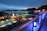 A nighttime scene overlooking a luxurious resort with multiple illuminated swimming pools. The view extends over a body of water with silhouetted islands in the distance. The area is adorned with palm trees and decorative lighting, creating a serene and inviting atmosphere.
