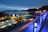 A nighttime scene overlooking a luxurious resort with multiple illuminated swimming pools. The view extends over a body of water with silhouetted islands in the distance. The area is adorned with palm trees and decorative lighting, creating a serene and inviting atmosphere.