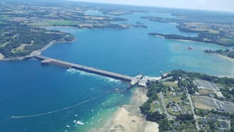 An aerial view of a coastal area with a prominent dam or bridge crossing a body of water. The landscape includes lush greenery, small boats scattered in the water, sandy beaches, and cultivated fields. The shoreline curves with dense vegetation and residential areas visible near the coast.