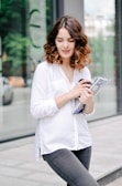 A young woman with wavy brown hair is standing outdoors, holding a rolled-up magazine and a phone in her hands. She is wearing a white blouse and dark gray jeans, and appears to be near a modern glass building with greenery reflected in the windows. The background includes a blurred view of a sidewalk and some parked vehicles.