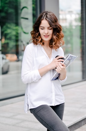 A young woman with wavy brown hair is standing outdoors, holding a rolled-up magazine and a phone in her hands. She is wearing a white blouse and dark gray jeans, and appears to be near a modern glass building with greenery reflected in the windows. The background includes a blurred view of a sidewalk and some parked vehicles.