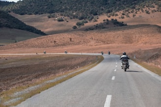 A group of motorcyclists riding along an empty road that winds through a hilly landscape. The terrain is mostly arid with patches of greenery and areas of sparse vegetation. The sky is clear, indicating a bright day.