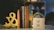 A collection of books is arranged vertically on a table, flanked by decorative bookends shaped like an ampersand. Next to the books, there is a white block with the inspirational quote 'WRITE WITHOUT FEAR. EDIT WITHOUT MERCY.' A pair of black glasses rests on the table in front.
