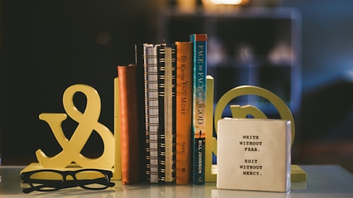 A collection of books is arranged vertically on a table, flanked by decorative bookends shaped like an ampersand. Next to the books, there is a white block with the inspirational quote 'WRITE WITHOUT FEAR. EDIT WITHOUT MERCY.' A pair of black glasses rests on the table in front.