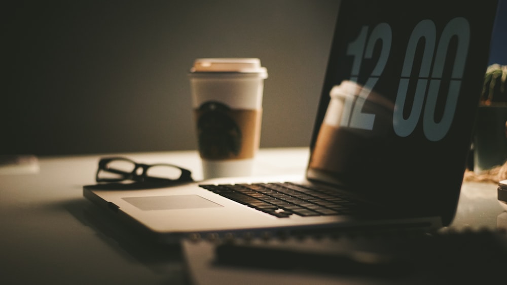 grey laptop computer displaying 12:00 on table near coffee cup and eyeglasses