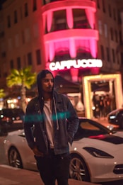 A person wearing a denim jacket and hoodie is standing near a white sports car on a dimly lit street. In the background, there's a building with neon pink lighting spelling 'CAPPUCCINO.' There are palm trees and other cars along the street, suggesting a nightlife scene.