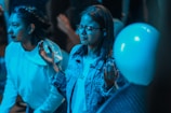 A group of people appears to be in a contemplative or attentive state. One individual, wearing glasses and a denim jacket, has both hands raised slightly, possibly in a gesture of thoughtfulness or engagement. Another person nearby is wearing a light hoodie. The lighting casts a blue tone over the scene, creating a calm or introspective ambiance. A balloon is visible in the foreground, hinting at a possible celebratory or casual environment.
