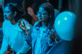 A group of people appears to be in a contemplative or attentive state. One individual, wearing glasses and a denim jacket, has both hands raised slightly, possibly in a gesture of thoughtfulness or engagement. Another person nearby is wearing a light hoodie. The lighting casts a blue tone over the scene, creating a calm or introspective ambiance. A balloon is visible in the foreground, hinting at a possible celebratory or casual environment.