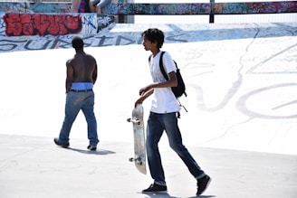 Two young individuals are at a skatepark. One is holding a skateboard and wearing a backpack, while the other is shirtless with blue jeans. Graffiti covers the surrounding walls, adding a vibrant backdrop.