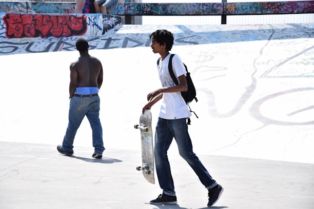 Two young individuals are at a skatepark. One is holding a skateboard and wearing a backpack, while the other is shirtless with blue jeans. Graffiti covers the surrounding walls, adding a vibrant backdrop.