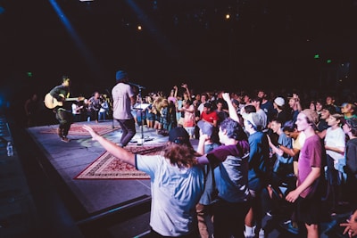 A lively music performance is happening on stage with a crowd of people enthusiastically enjoying the show. The audience is standing close to the stage, with many participants dancing and cheering. A musician is playing a guitar while another artist is singing into a microphone. The environment is dimly lit with colorful stage lights enhancing the atmosphere.