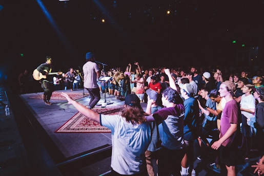 A lively music performance is happening on stage with a crowd of people enthusiastically enjoying the show. The audience is standing close to the stage, with many participants dancing and cheering. A musician is playing a guitar while another artist is singing into a microphone. The environment is dimly lit with colorful stage lights enhancing the atmosphere.