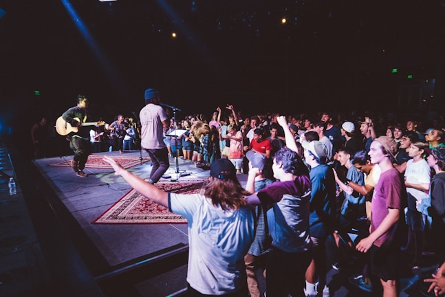 A lively music performance is happening on stage with a crowd of people enthusiastically enjoying the show. The audience is standing close to the stage, with many participants dancing and cheering. A musician is playing a guitar while another artist is singing into a microphone. The environment is dimly lit with colorful stage lights enhancing the atmosphere.