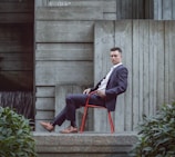 A man in a suit sits on a red chair, leaning back against a concrete wall with modern, textured surfaces. Green foliage is visible in the foreground, contrasting with the stark industrial backdrop.