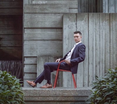 A man in a suit sits on a red chair, leaning back against a concrete wall with modern, textured surfaces. Green foliage is visible in the foreground, contrasting with the stark industrial backdrop.
