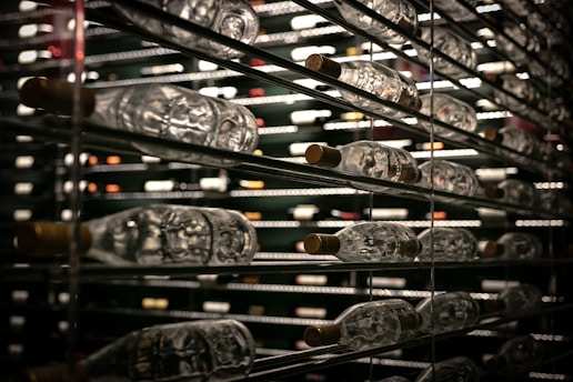 Rows of wine bottles are stored horizontally on metal shelves. The reflective glass and dim lighting create a luxurious atmosphere, with a focus on the variety and quantity of bottles.