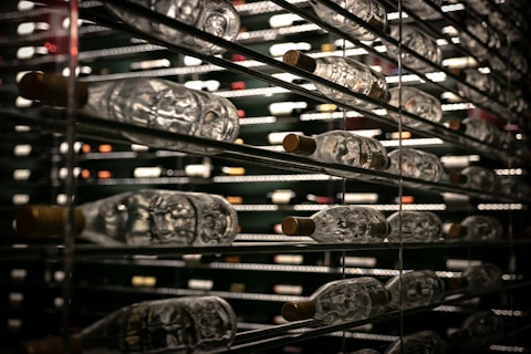 Rows of wine bottles are stored horizontally on metal shelves. The reflective glass and dim lighting create a luxurious atmosphere, with a focus on the variety and quantity of bottles.