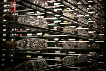 Rows of wine bottles are stored horizontally on metal shelves. The reflective glass and dim lighting create a luxurious atmosphere, with a focus on the variety and quantity of bottles.