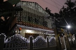 A two-story house adorned with decorative string lights that illuminate its facade, creating a festive atmosphere. The lights are draped along the railings and entrance gate, emitting a warm glow. The house is situated in a lush, leafy setting with various trees visible in the background. The lighting and colors suggest a celebratory occasion during nighttime.