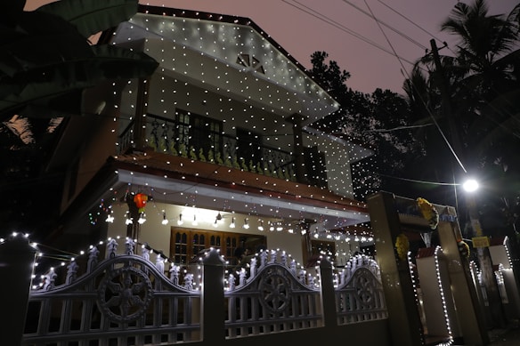 A two-story house adorned with decorative string lights that illuminate its facade, creating a festive atmosphere. The lights are draped along the railings and entrance gate, emitting a warm glow. The house is situated in a lush, leafy setting with various trees visible in the background. The lighting and colors suggest a celebratory occasion during nighttime.