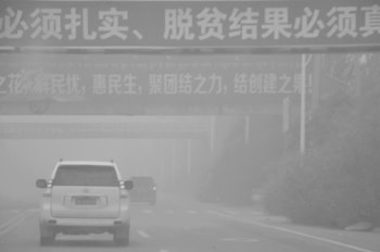 A foggy road scene with several vehicles driving. Overhead, multiple signs with Chinese characters are visible, partially obscured by the fog. The road is lined with trees and vegetation, also hazy due to the weather conditions.