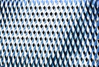 A facade of a building is covered with a repetitive pattern of geometric shapes. Each element consists of diamond shapes and triangular facets, creating an illusion of texture and depth with reflective surfaces. The elements are predominantly in shades of blue and white.