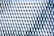 A facade of a building is covered with a repetitive pattern of geometric shapes. Each element consists of diamond shapes and triangular facets, creating an illusion of texture and depth with reflective surfaces. The elements are predominantly in shades of blue and white.