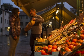 A man is handing a bunch of bananas to a woman at an outdoor market. The market stall is filled with an array of fruits, such as oranges and bananas, neatly arranged in boxes. The scene takes place in the evening under the warm glow of lights from the market stalls, with a backdrop of old buildings and a wet street surface.