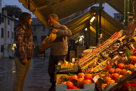 A man is handing a bunch of bananas to a woman at an outdoor market. The market stall is filled with an array of fruits, such as oranges and bananas, neatly arranged in boxes. The scene takes place in the evening under the warm glow of lights from the market stalls, with a backdrop of old buildings and a wet street surface.